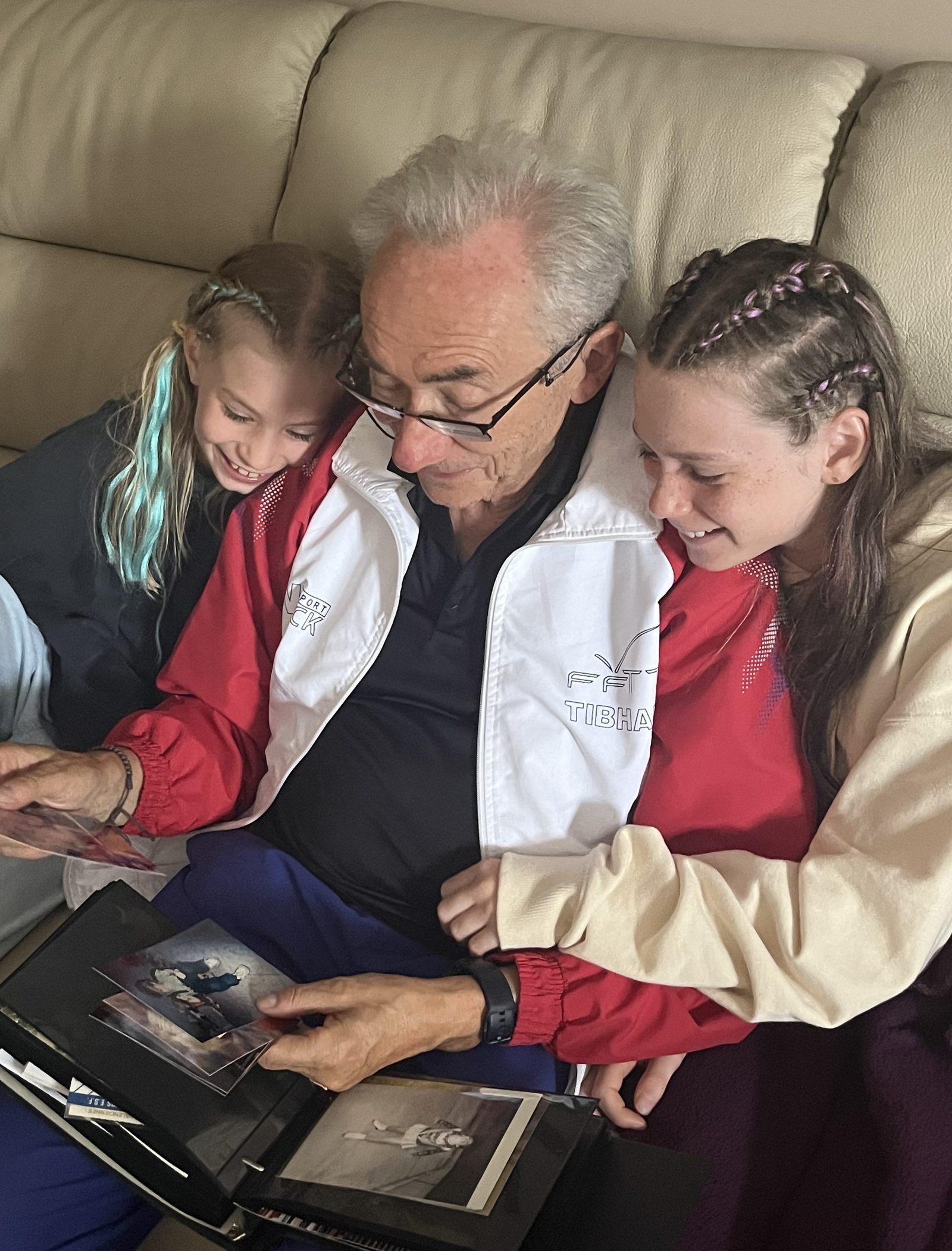Un grand-père feuillette un album photo avec ses deux petites-filles, blotties contre lui sur un canapé.